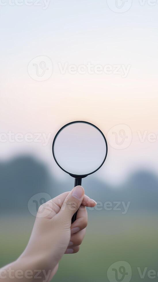 Hand holding a magnifying glass against a soft pastel sky, with blurred greenery in the background, symbolizing exploration, curiosity, and discovery in nature photo