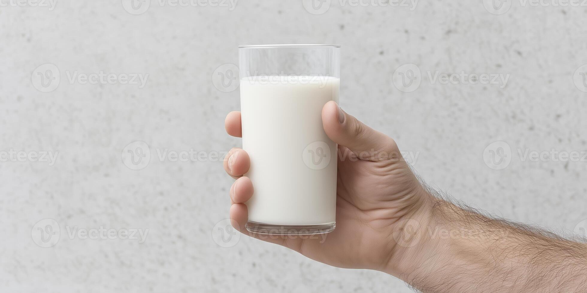 Hand holding a clear glass filled with fresh milk against a textured gray background, showcasing the beverage's creamy texture and inviting appearance for culinary or health-related themes photo