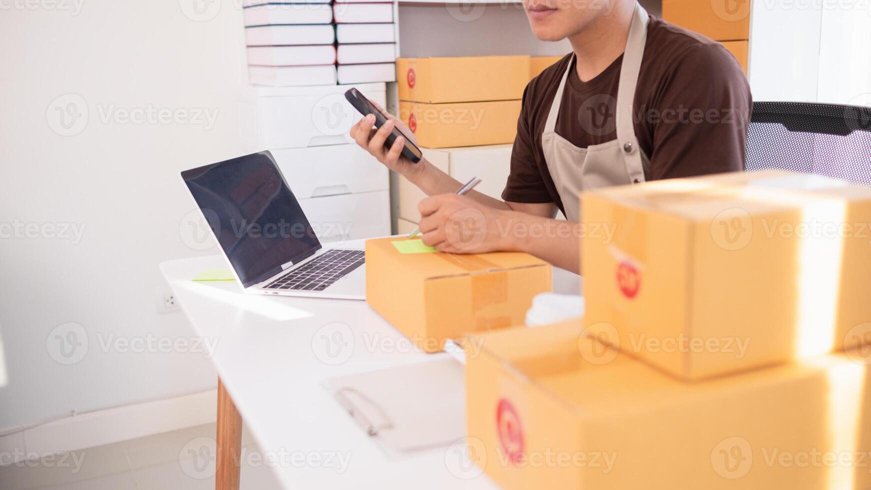 A man in an apron is using a cell phone while sitting at a desk with boxes photo