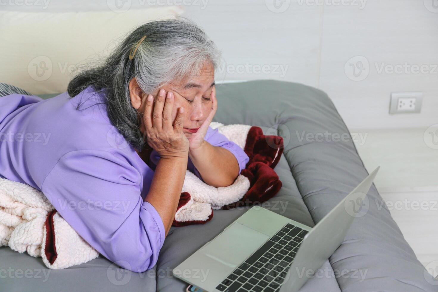 Exhausted Senior Woman Resting While Using Laptop, Mature Woman with Fatigue Lying on Bed with Computer, Tired Elderly Lady Propping Her Head on Hands Looking at Screen photo