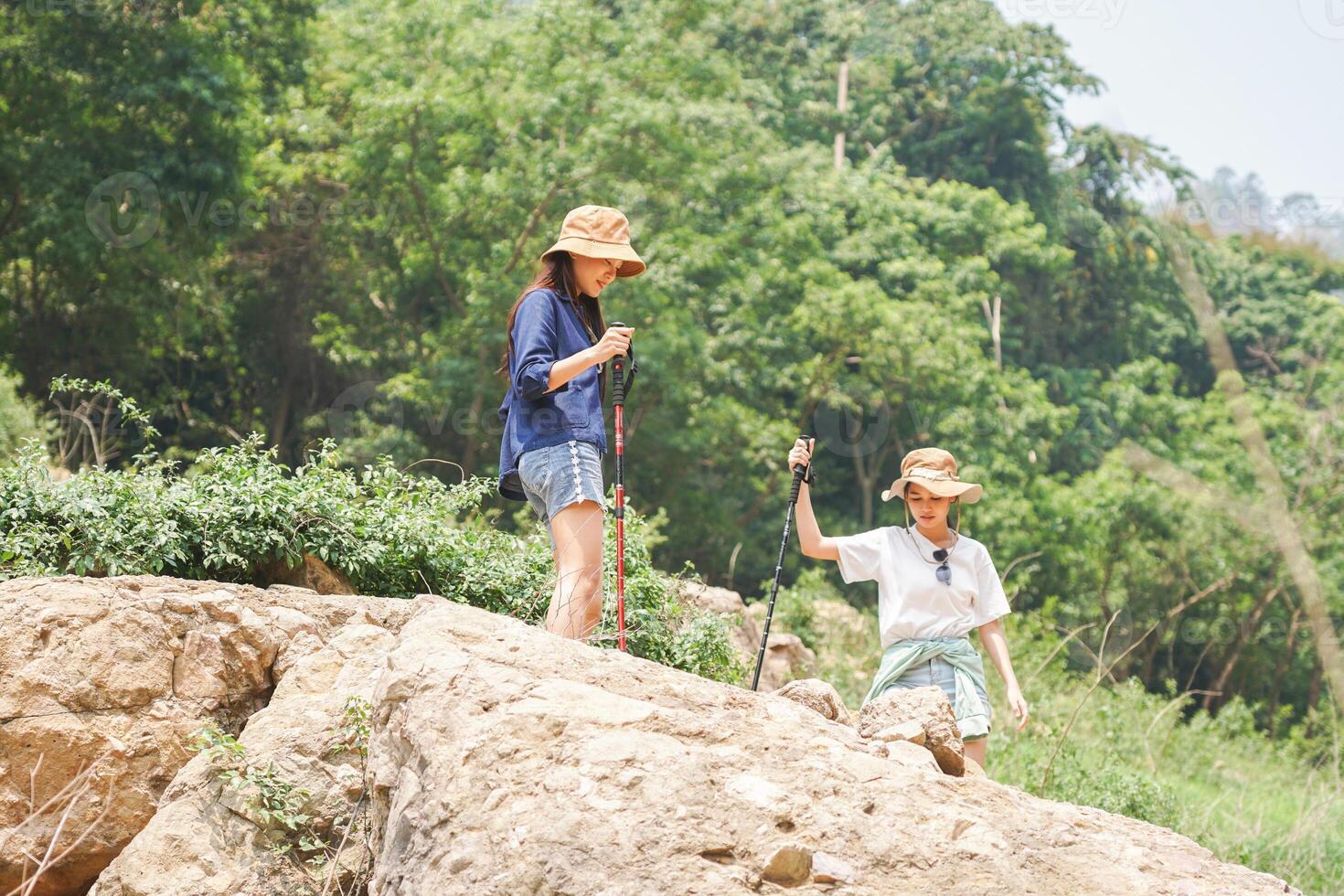 Friends Exploring Rocky Terrain with Trekking Poles, Young Asian Women Hiking in Lush Green Mountains, Female Hikers on a Sunny Mountain Trail Enjoying Nature photo