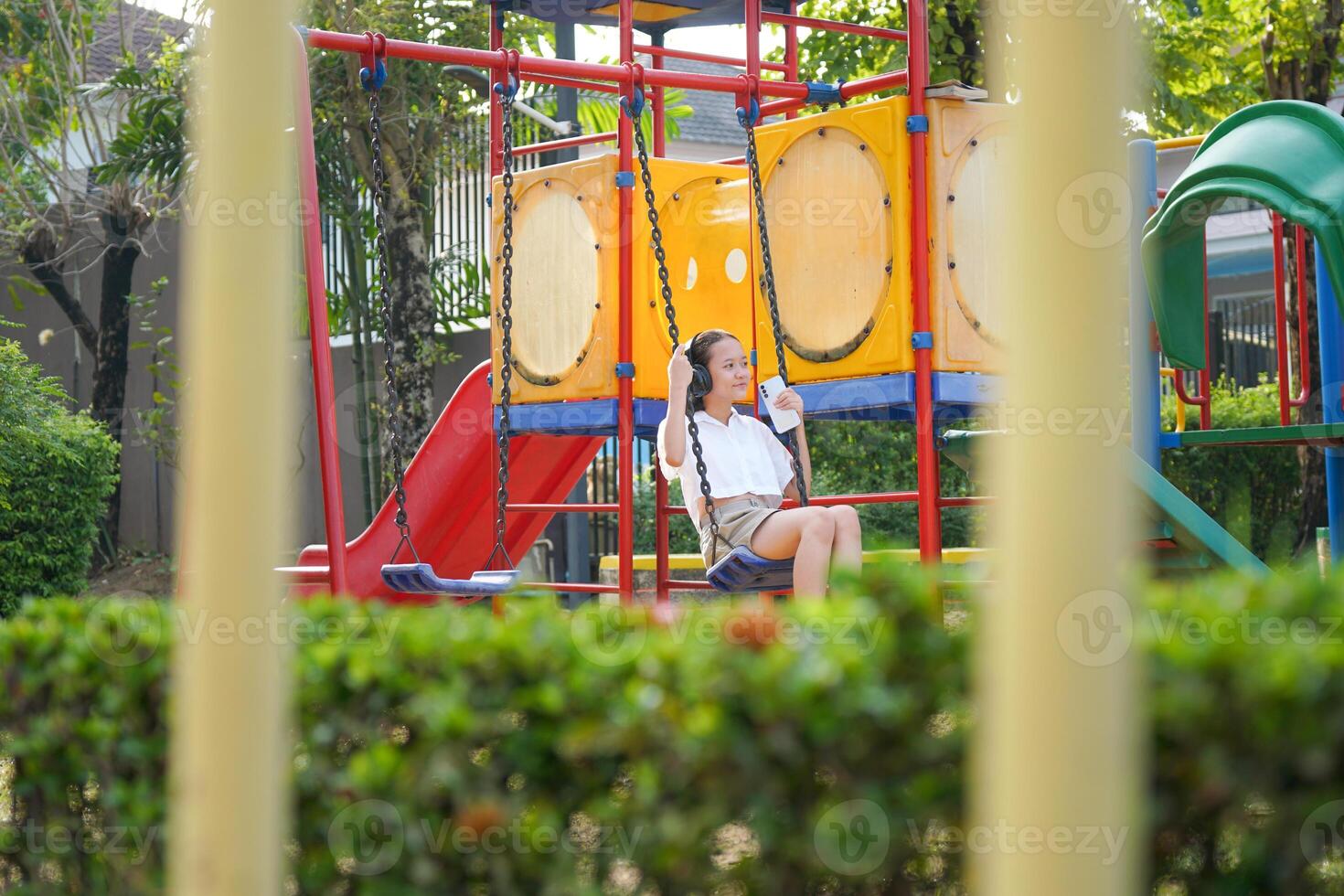 Young Asian Girl Enjoys Music on a Swing at a Colorful Playground, Happy Child Relaxing with Headphones on a Swing in a Park photo