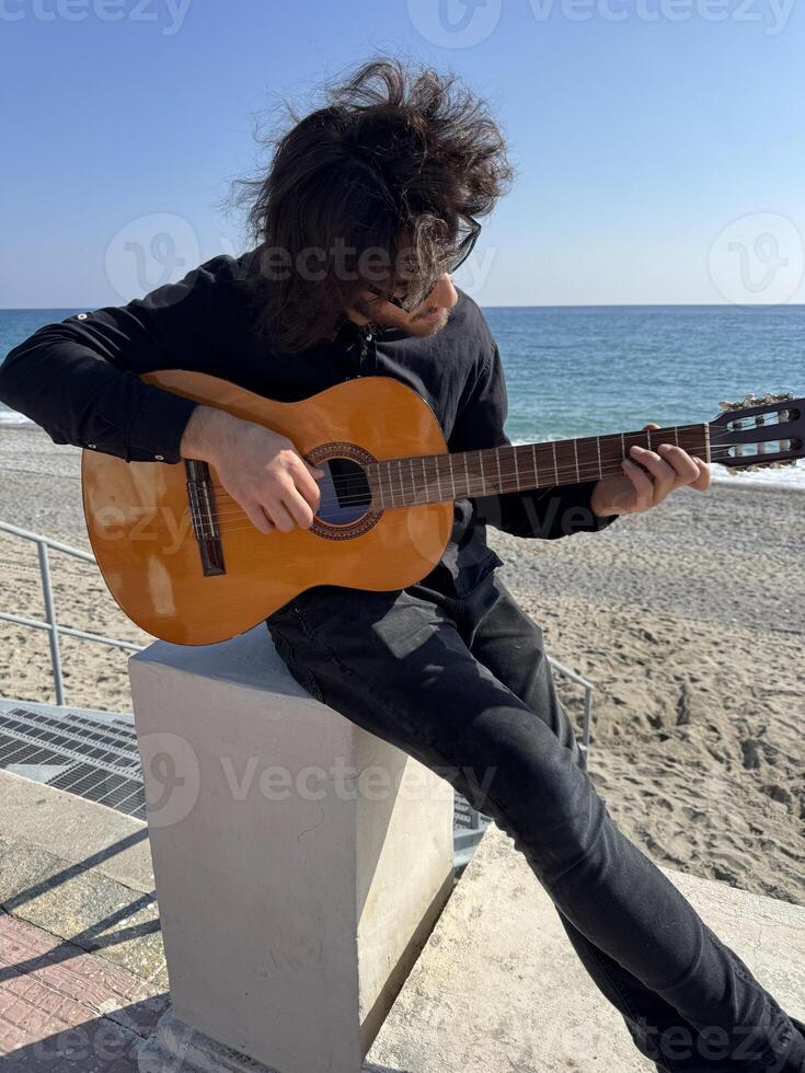 Long Hair Boy Plays Classical Guitar by the Ocean in Summer Dressed in Outdoor photo