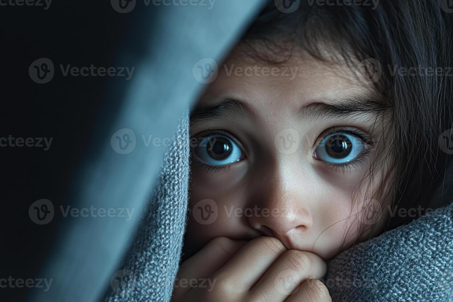 Child expresses fear and anxiety while hiding under a blanket in dimly lit room photo