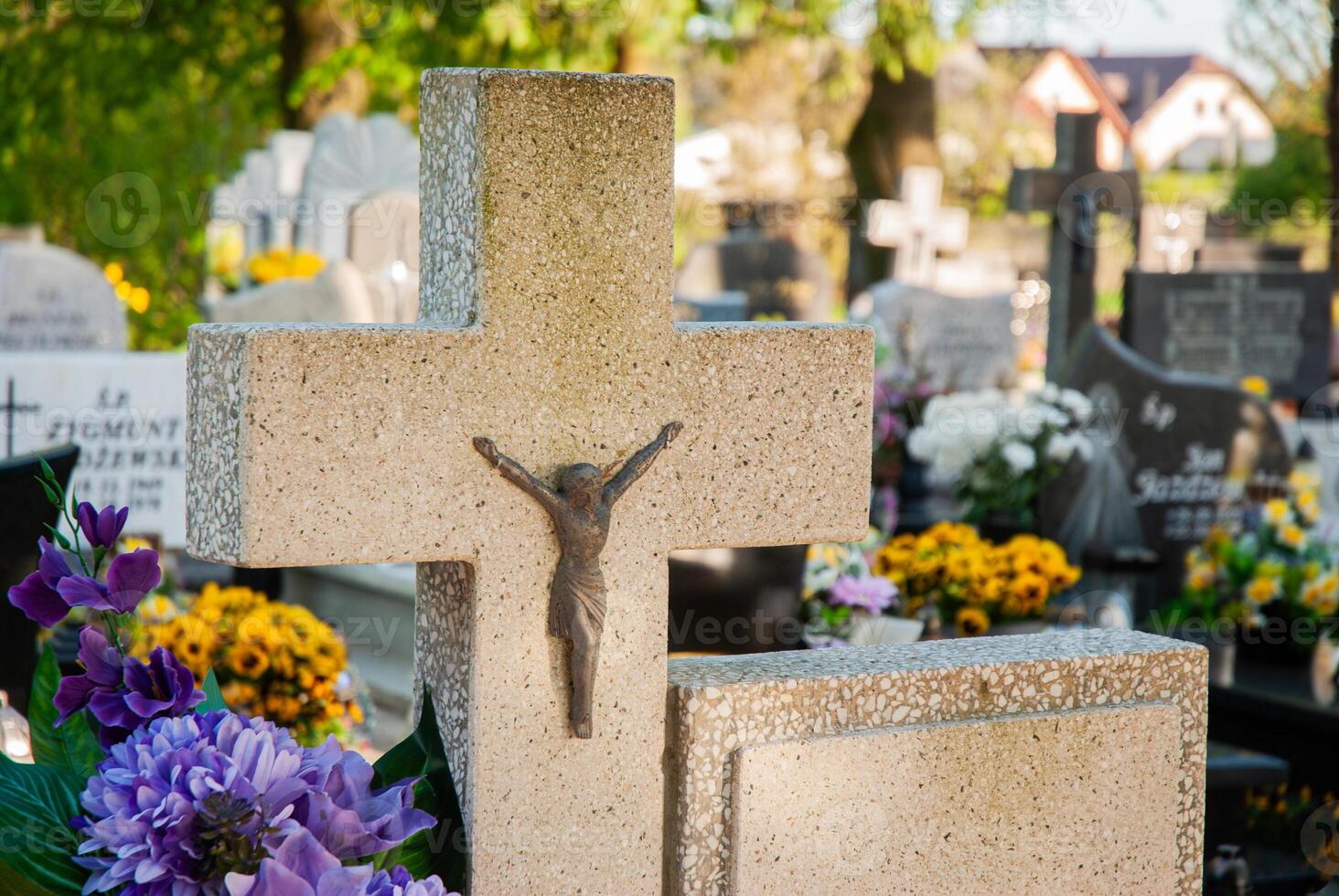 Stone cross adorned with a crucifix stands among vibrant flowers and gravestones in a peaceful cemetery under a sunny, clear sky photo
