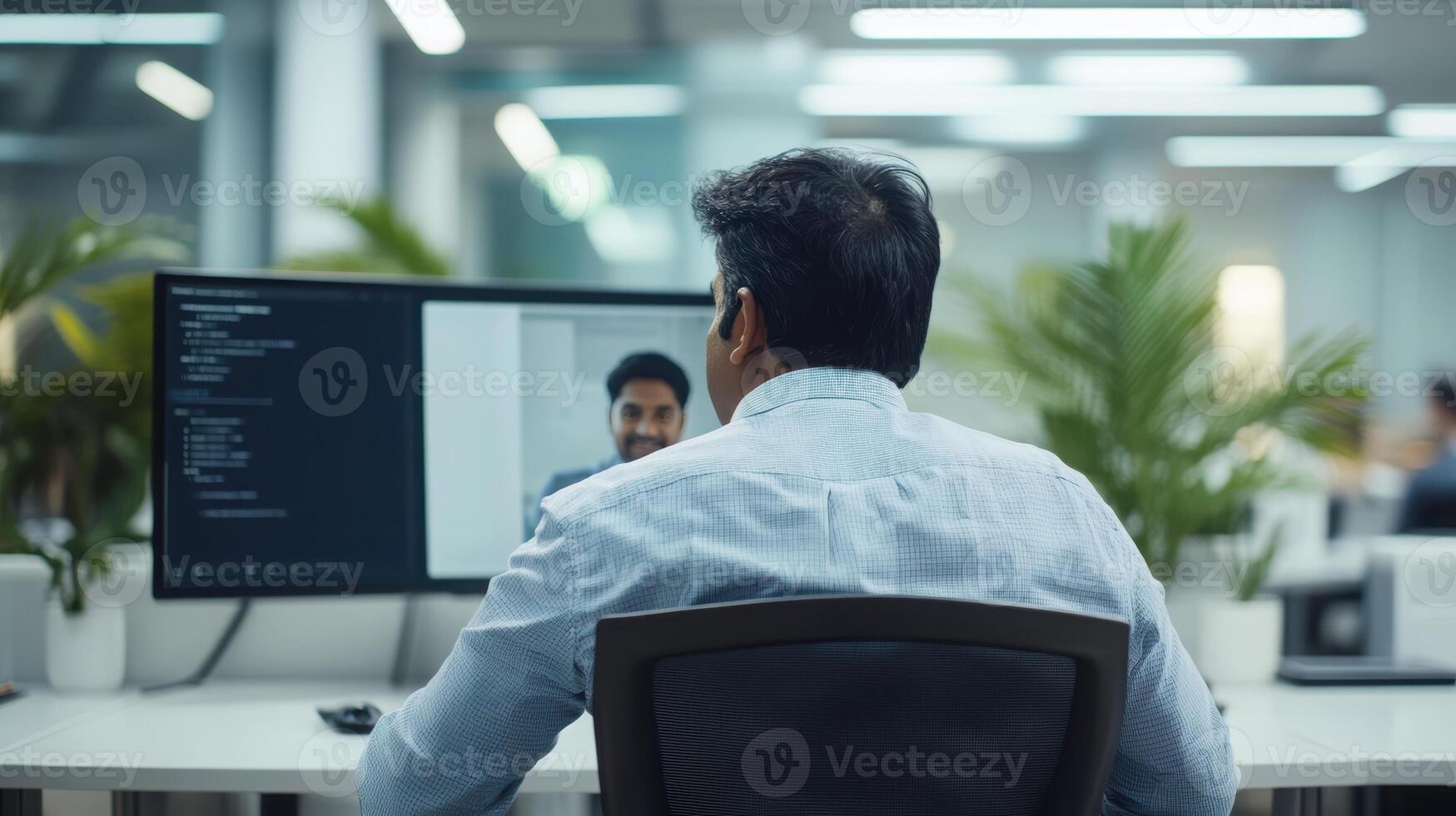 Indian male adult in modern office working on computer while conferencing with colleague, surrounded by plants and bright lighting. Mentor worker photo