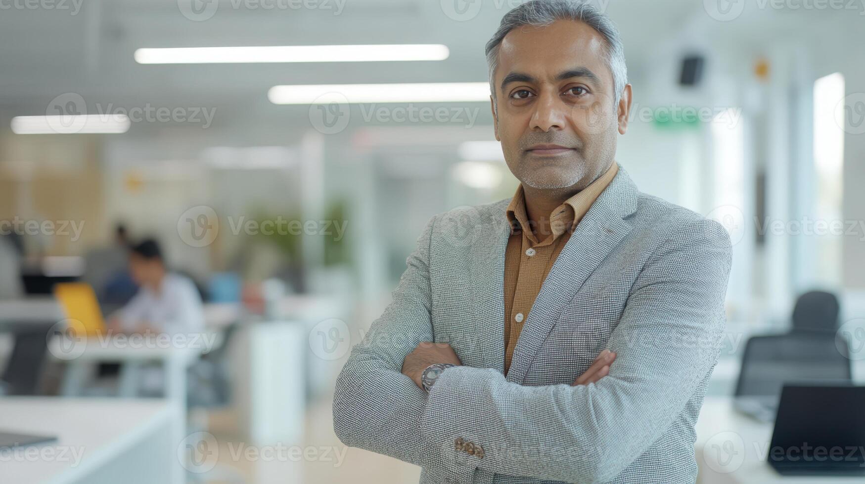 Confident mature indian male in business attire standing with arms crossed in modern open office space with blurred background. Mentor worker photo