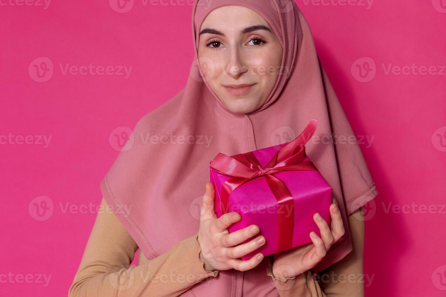 An Arab female teen in a pink hijab holding a gift. A girl in Muslim religious clothes smiles against a bright background photo