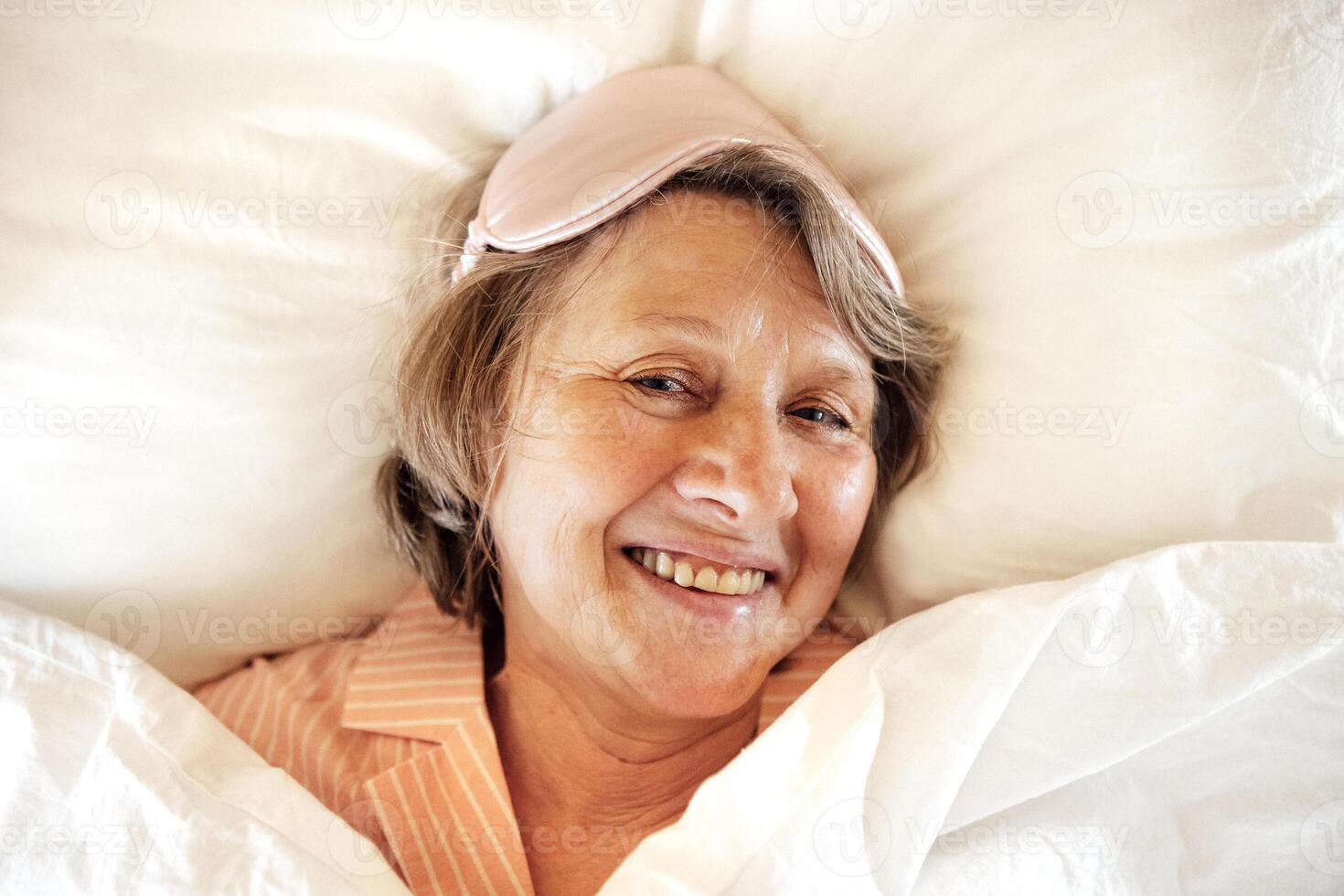 Portrait of an elderly woman lying in bed and smiling. A mature lady in pajamas and a sleep mask wakes up and laughs. photo