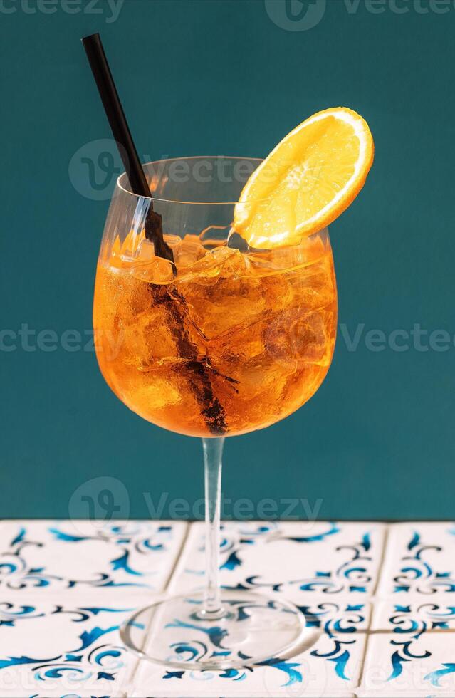 A glass with aperol spritz and a piece of lemon. A goblet of cool delicious orange drink with ice cubes on blue background. Citrus alcoholic cocktail. photo