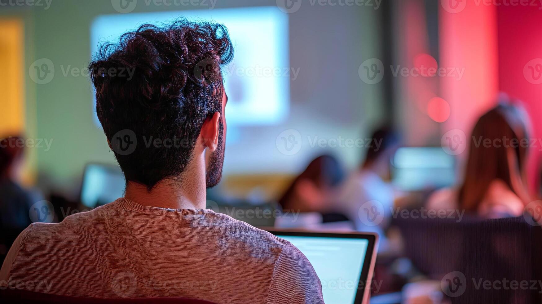 Young man with curly hair is focused on laptop during a seminar in a modern classroom, with blurred audience and colorful lighting creating an engaging atmosphere photo