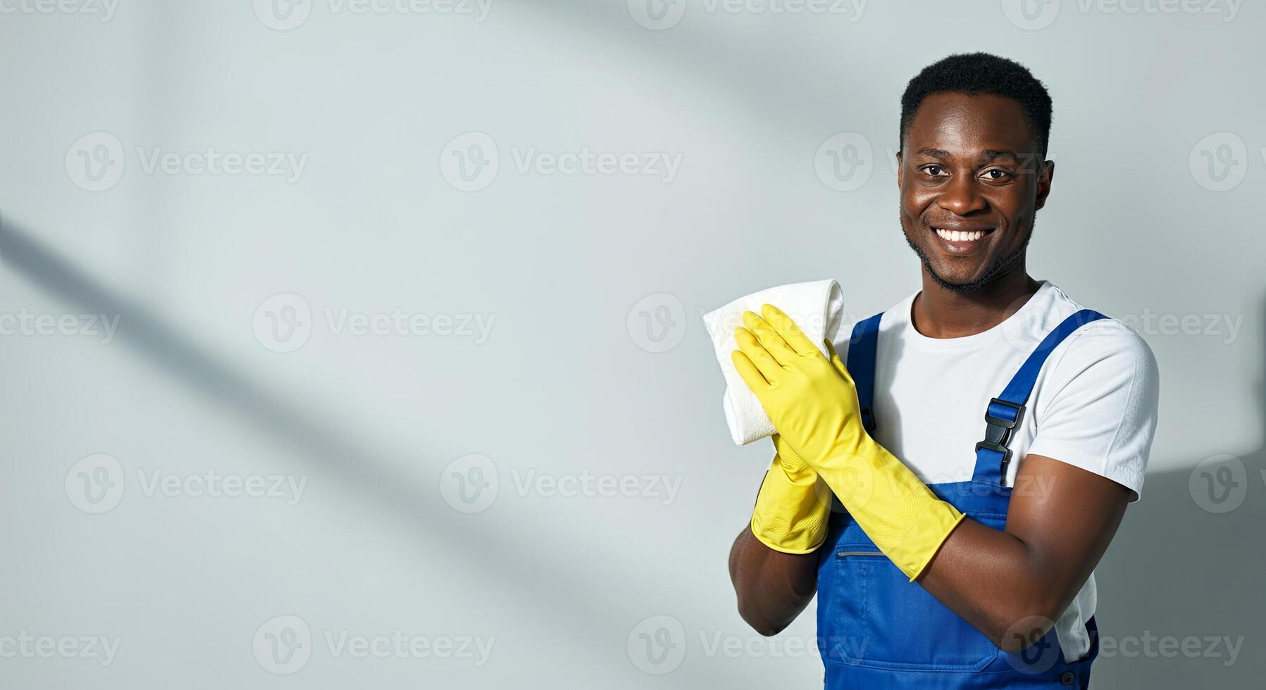 Smiling African American man in blue overalls and yellow gloves holds a cleaning cloth, showcasing a cheerful attitude while preparing to clean in a bright, minimalistic environment photo