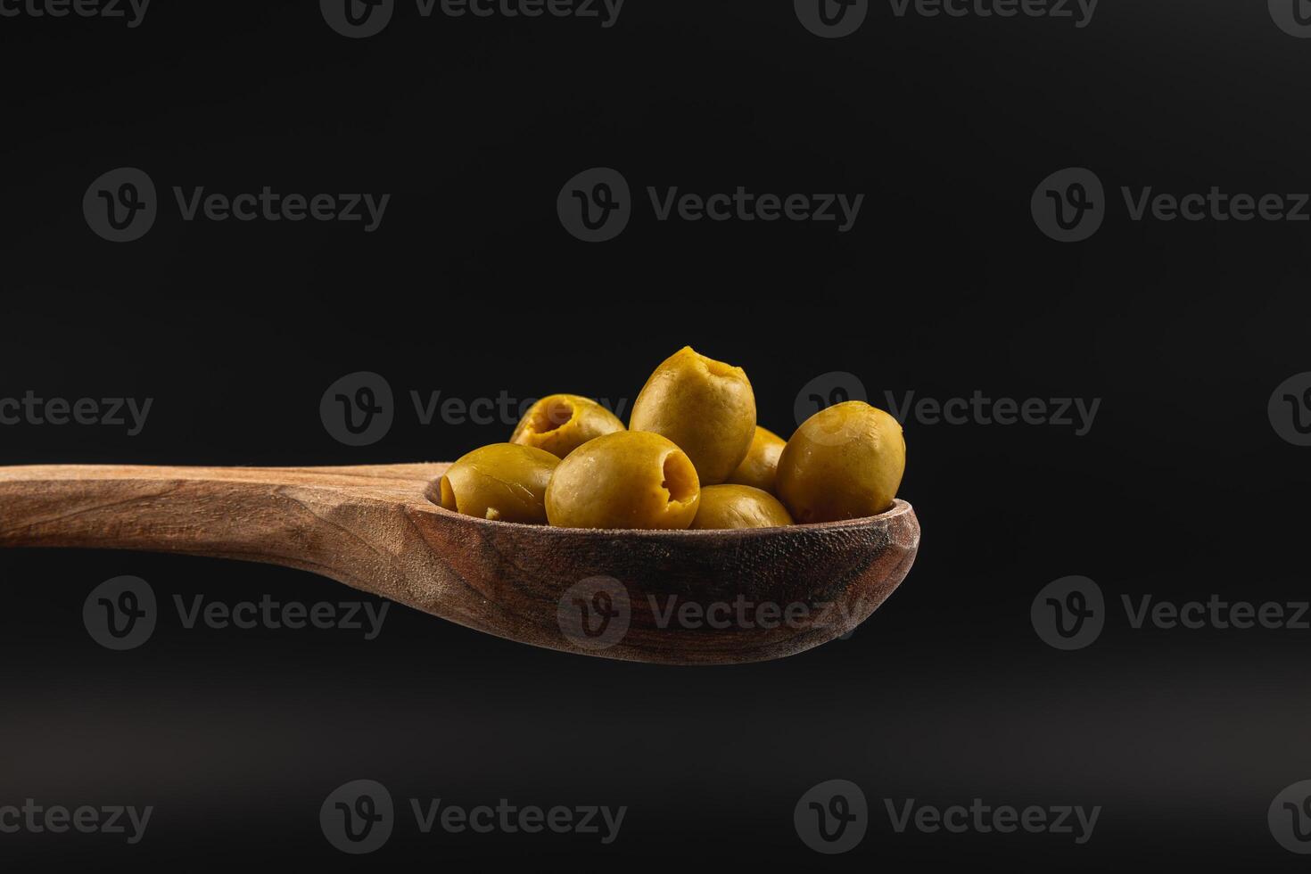Small green olives placed on a wooden spoon against a dark background photo