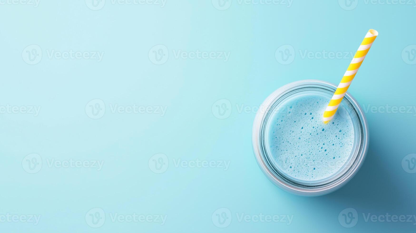 glass jar with yellow and white striped straw. The jar is filled with a blue smoothie or milkshake. The background is a light blue color photo