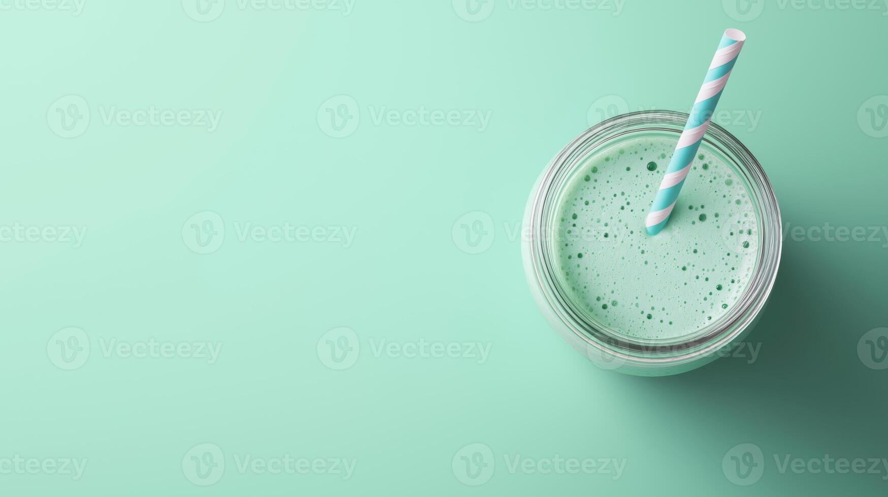 glass jar with blue and white striped straw. The jar is filled with a green smoothie or milkshake. The background is a light green color photo