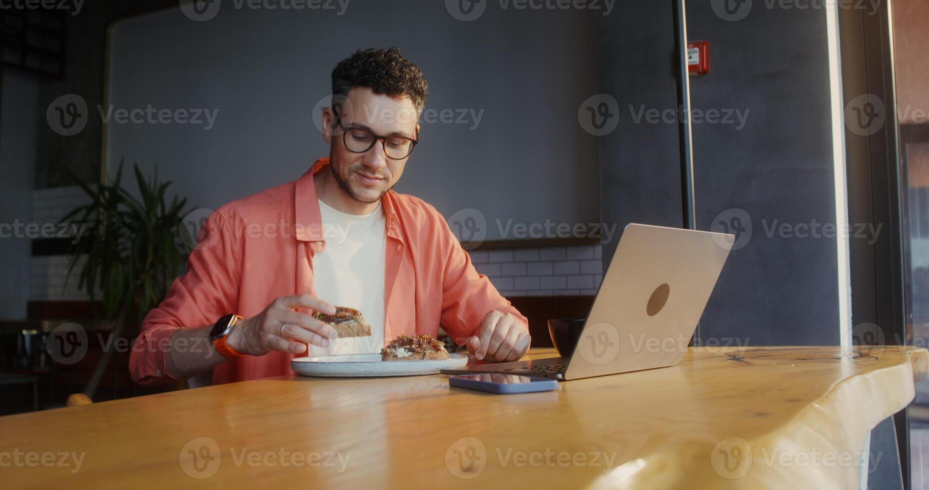 A stylish young man eats bruschetta and drinks coffee sitting in front of an open laptop in a cafe photo