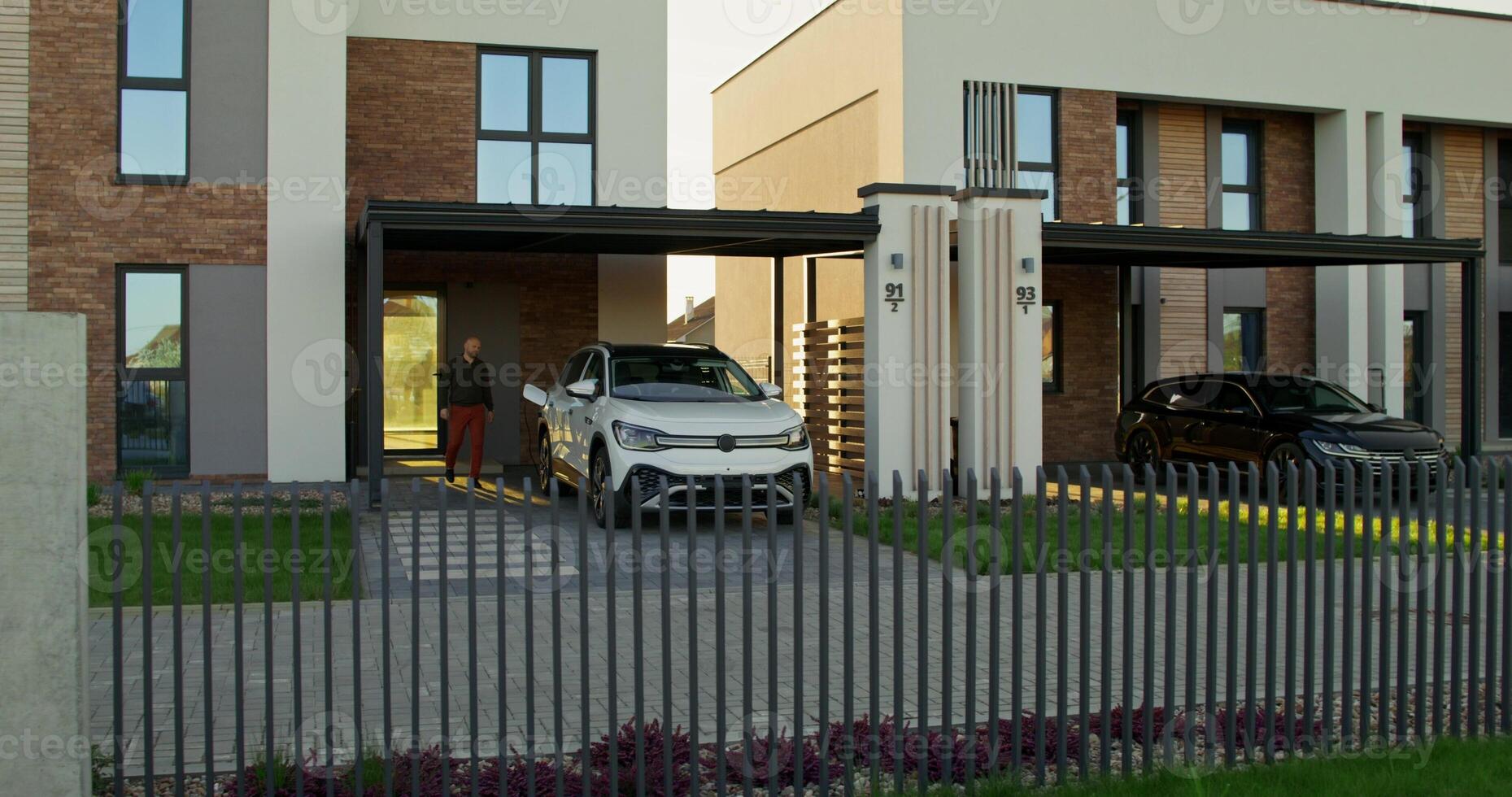 A man in casual clothes disconnects charging from an electric car parked in front of a stylish cottage. A sunny day photo