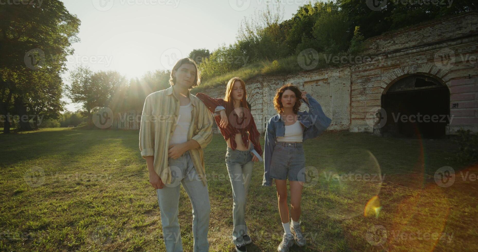 Three young friends look into the camera with a calm look standing near the ruins of a historic building. The fortress walls are overgrown with green vegetation. A sunny summer day photo