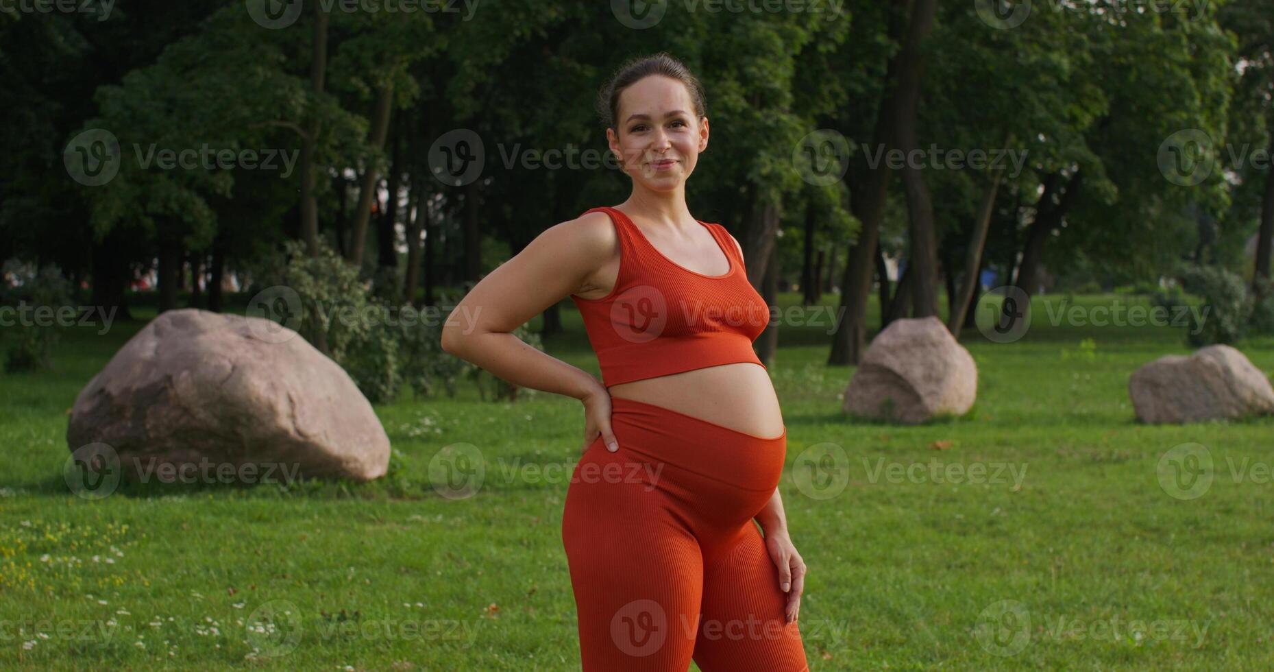 A pregnant woman in sports clothes smiling and looking at the camera standing in a city park on a summer day while doing sports photo