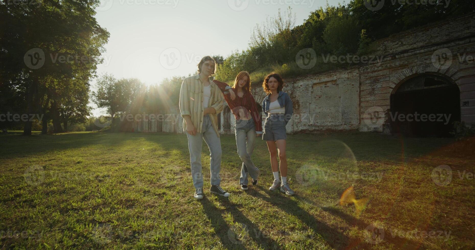 Three young friends look into the camera with a calm look standing near the ruins of a historic building. The fortress walls are overgrown with vegetation. photo