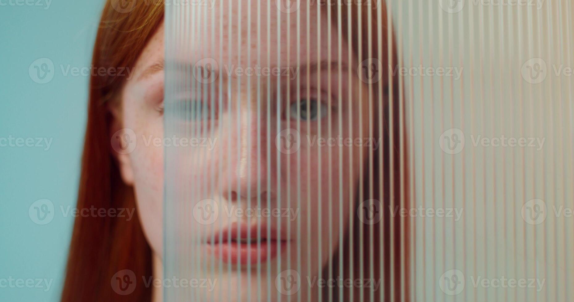 A red-haired woman with freckles is smiling at the camera while standing on a studio background. She hides her face behind plexiglass photo