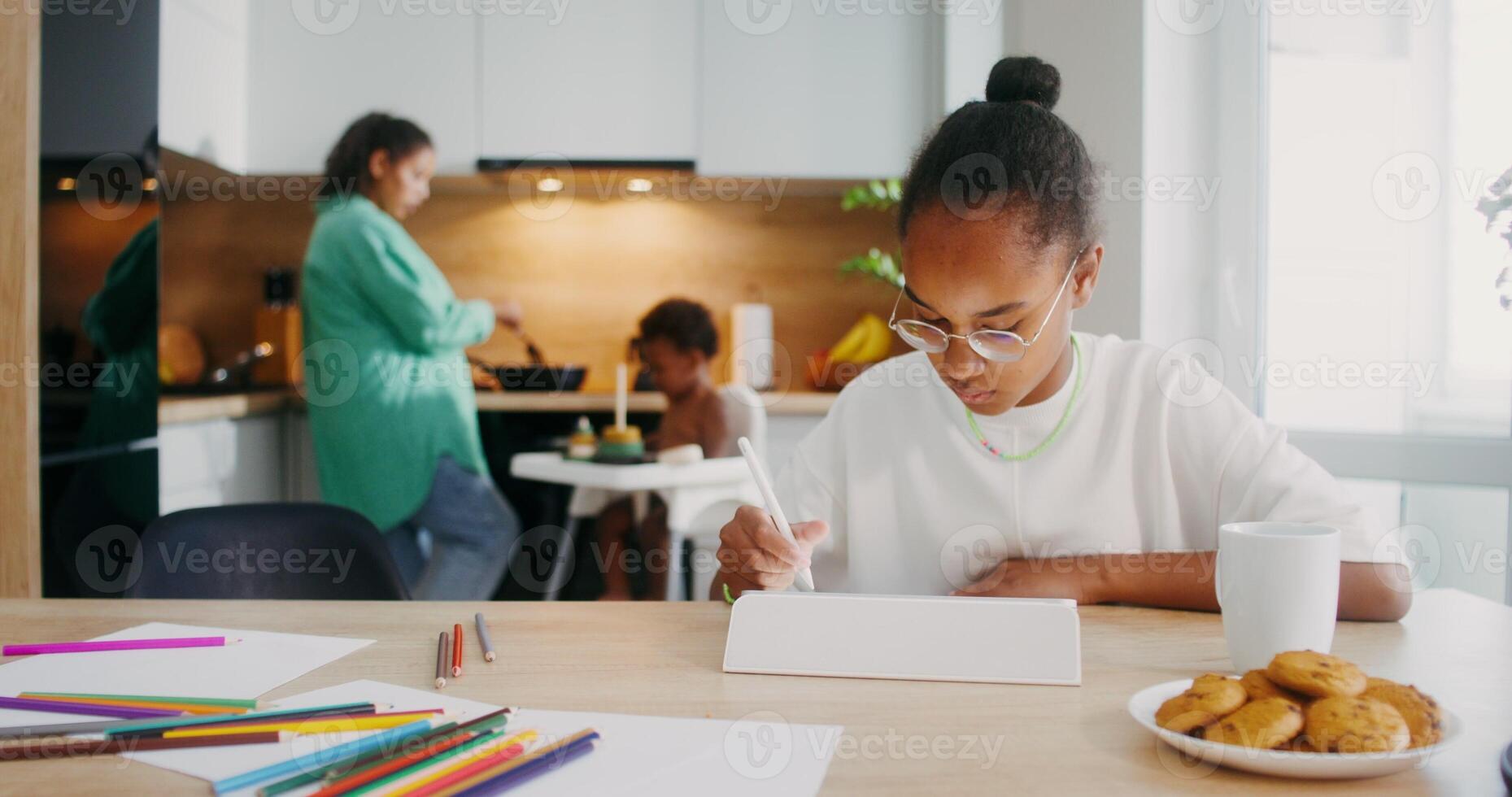 A girl draws with a stylus on a tablet while sitting at the kitchen table at home. Her family in the background is out of focus - sitting photo