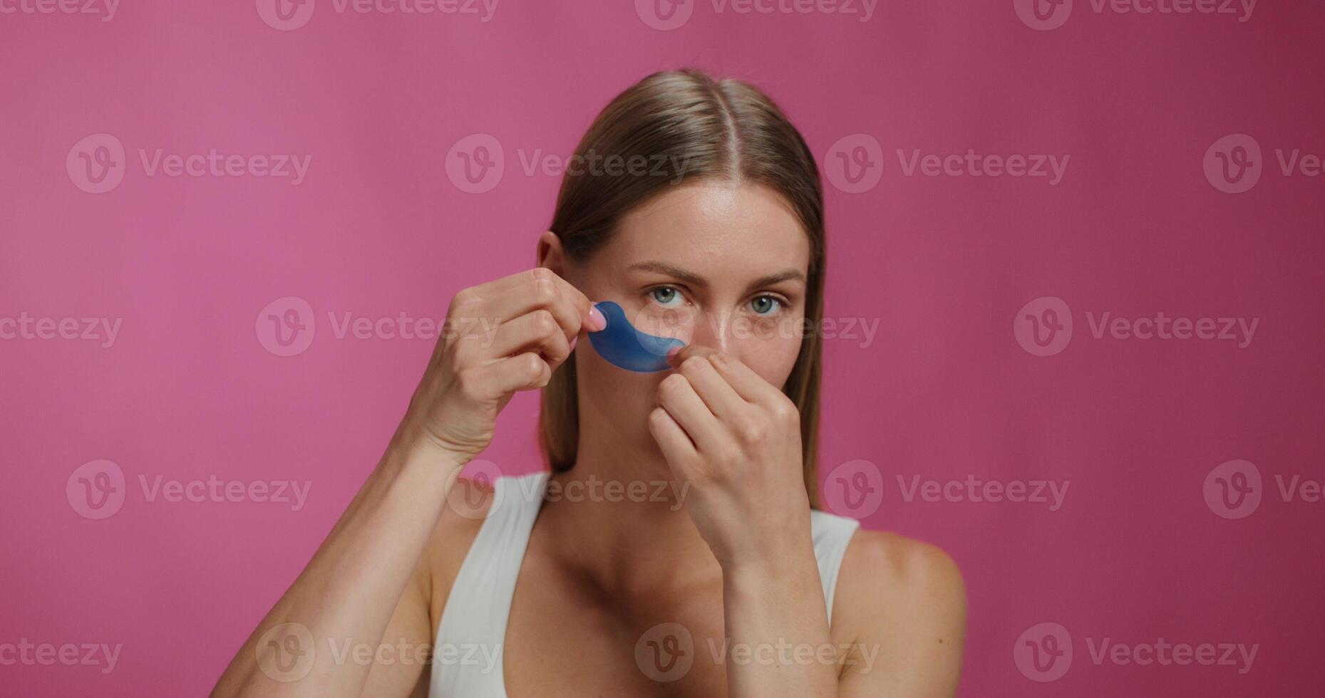 A woman applies patches to her lower eyelids. She smiles at the camera while standing on a pink background in the studio photo