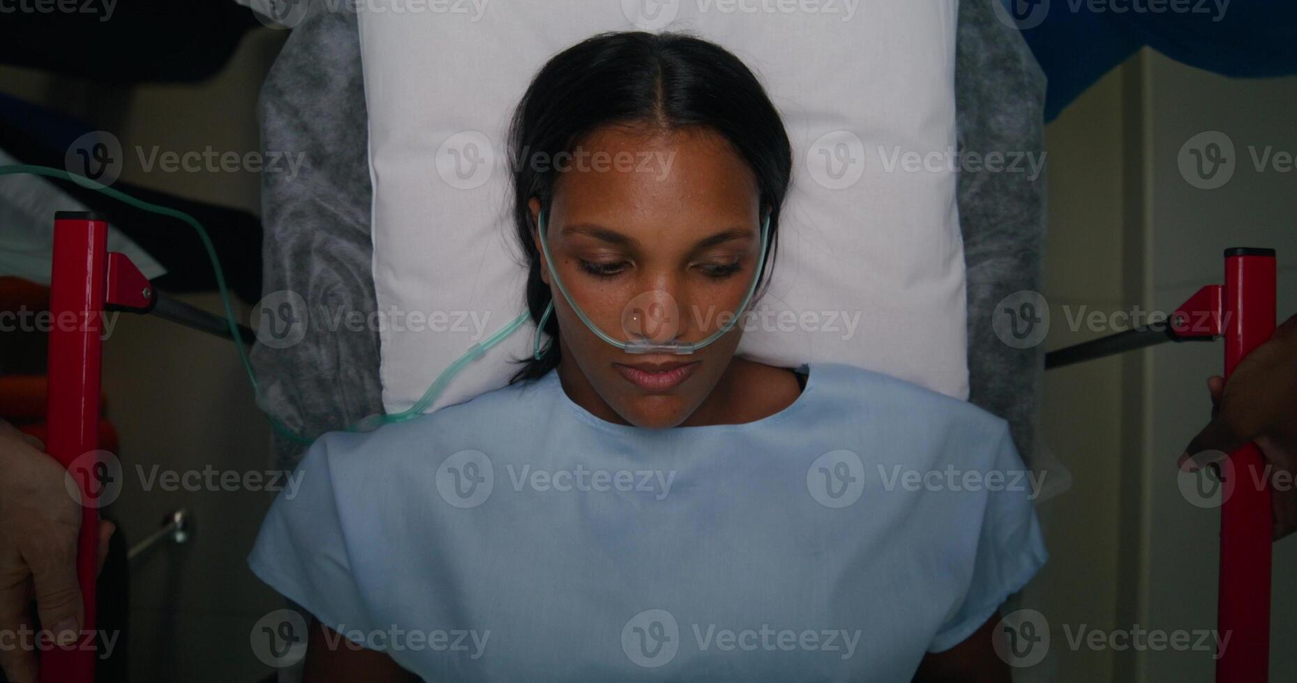 A woman with a breathing tube in her nose is lying on a gurney, top view. She is being driven down the hospital corridor photo