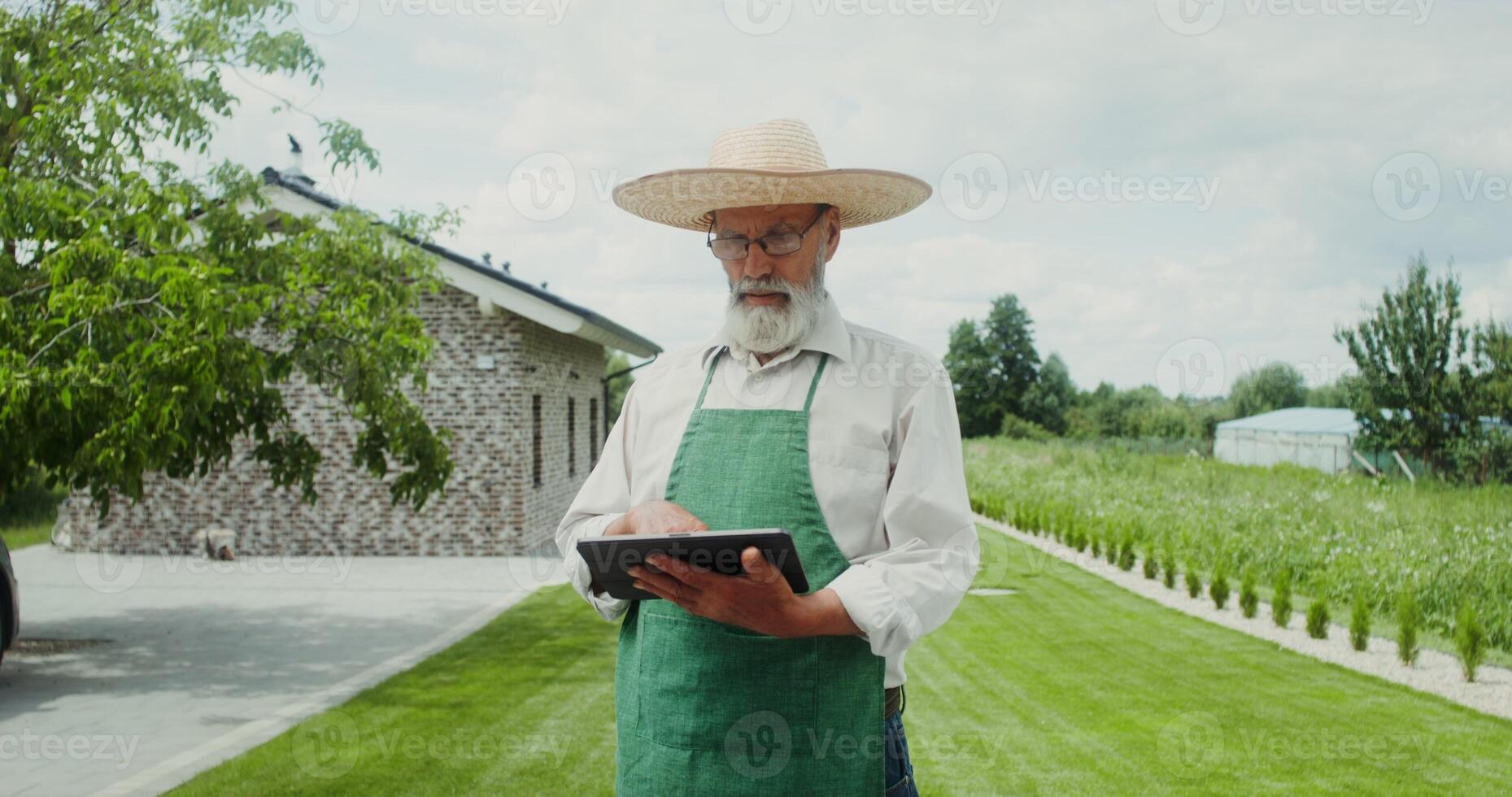 An elderly man with a gray beard, dressed in an apron and hat, smiles while looking at the camera standing in the backyard with a tablet in his hands photo