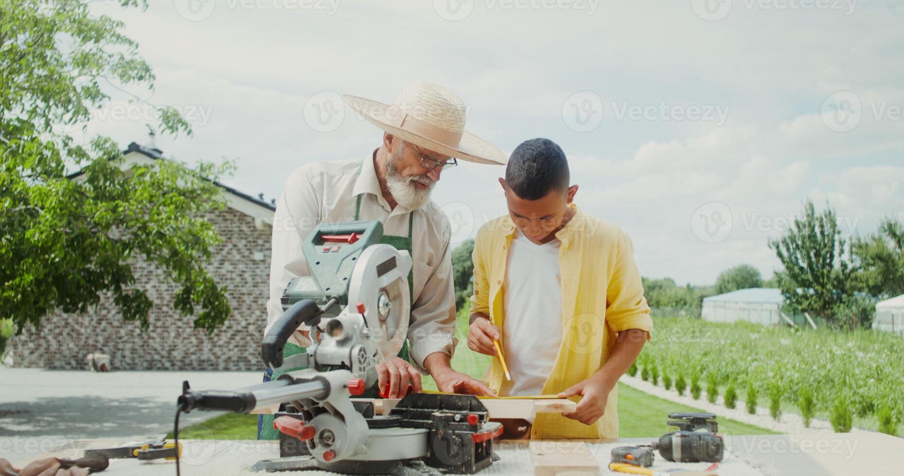 An elderly man teaches his grandson how to use a miter saw while standing in the backyard of his house - standing photo