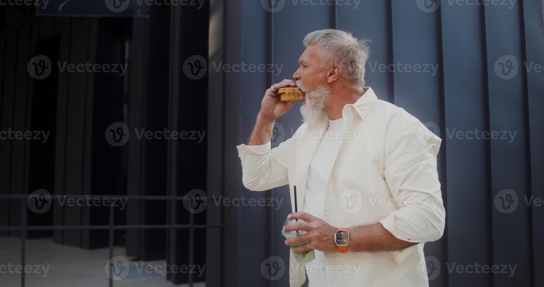 A mature gray-haired man with a mustache and beard eats a sandwich, and drinks lemonade while walking along a city street on a summer day photo