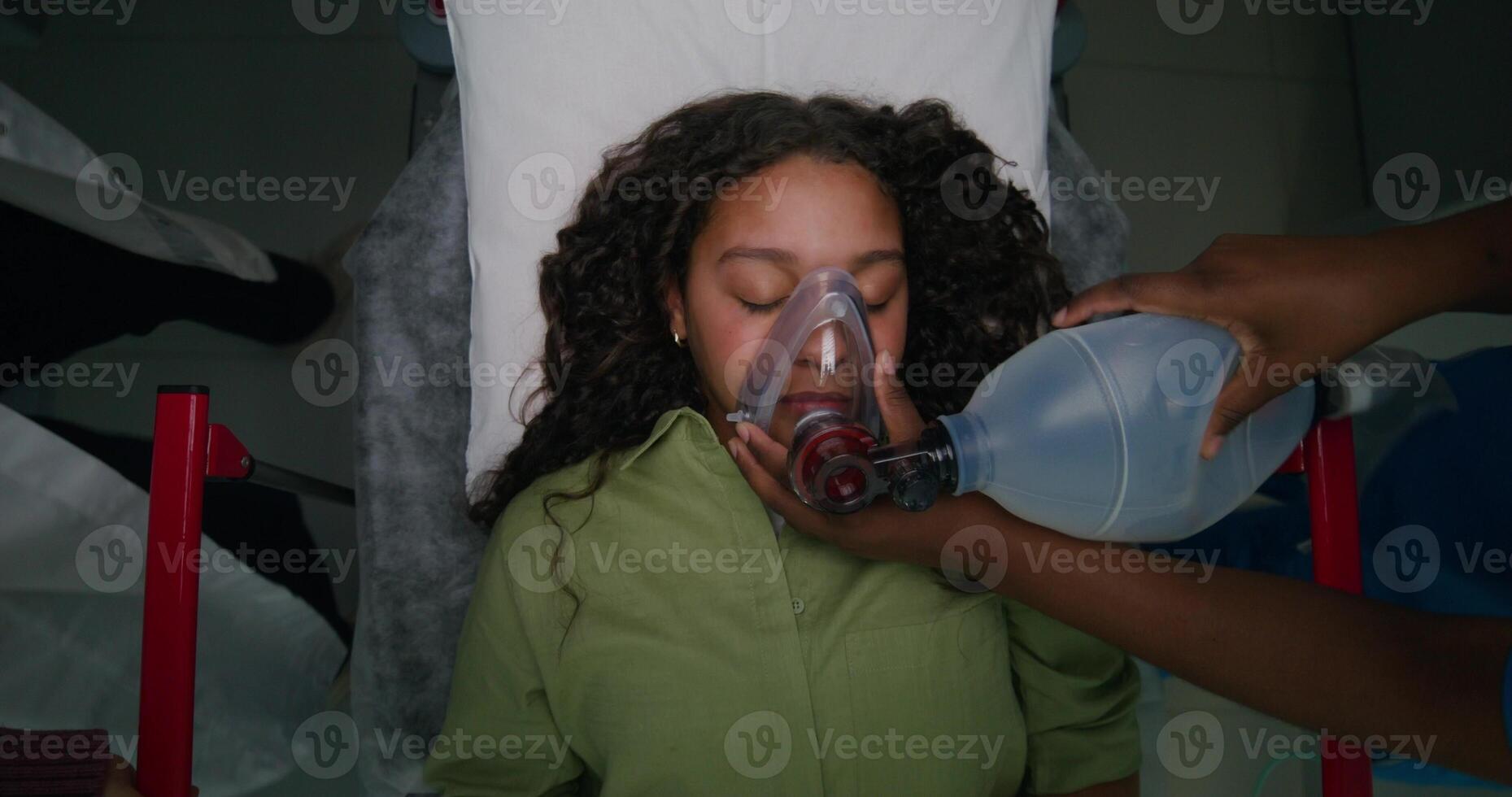 The patient is lying on a gurney with her eyes closed. The nurse gives her air through an oxygen mask. View from above. Emergency resuscitation photo