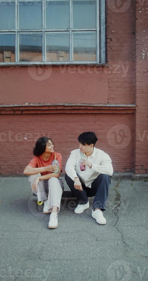 A young man and a woman are talking and drinking lemonade while sitting on a skateboard on a city street - skateboard photo