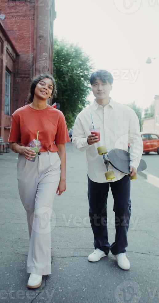 A young man and a woman smile, talk and drink lemonade while walking around the city. A man holds a longboard in his hands - longboard photo