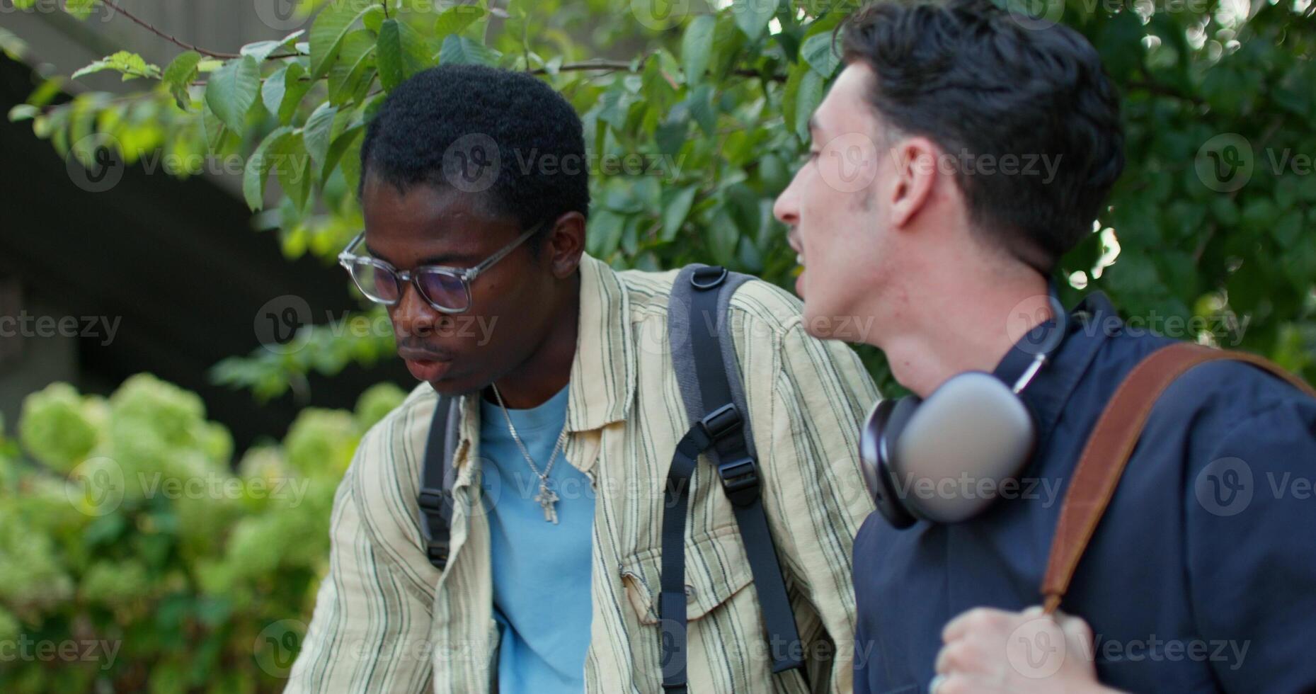 Two young men with backpacks on their shoulders are talking while sitting outdoors. photo
