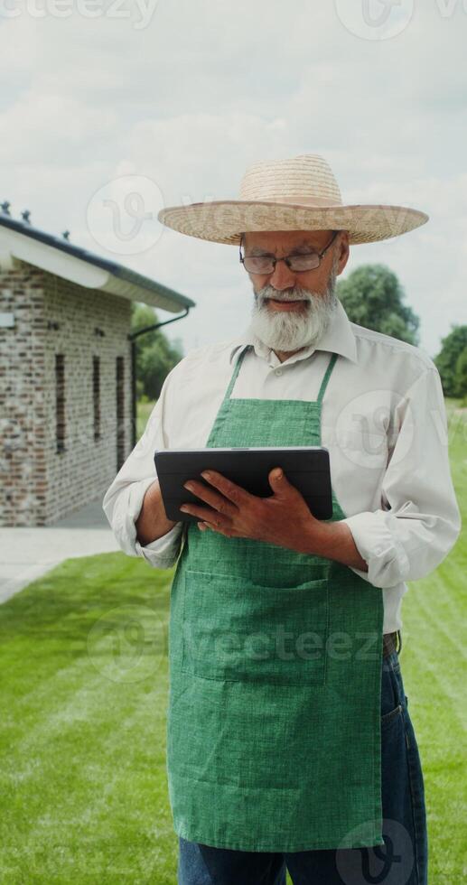 An elderly man with a gray beard, dressed in an apron and hat, smiles while looking at the camera standing in the backyard with a tablet in his hands - looking photo