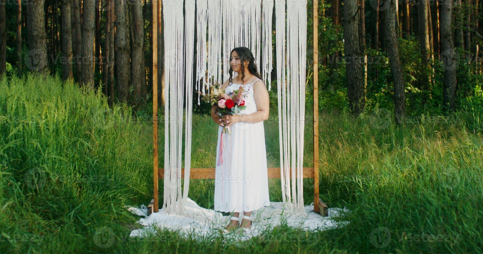 An African-American bride with a bouquet in her hands stands at a decorated arch in the nature photo