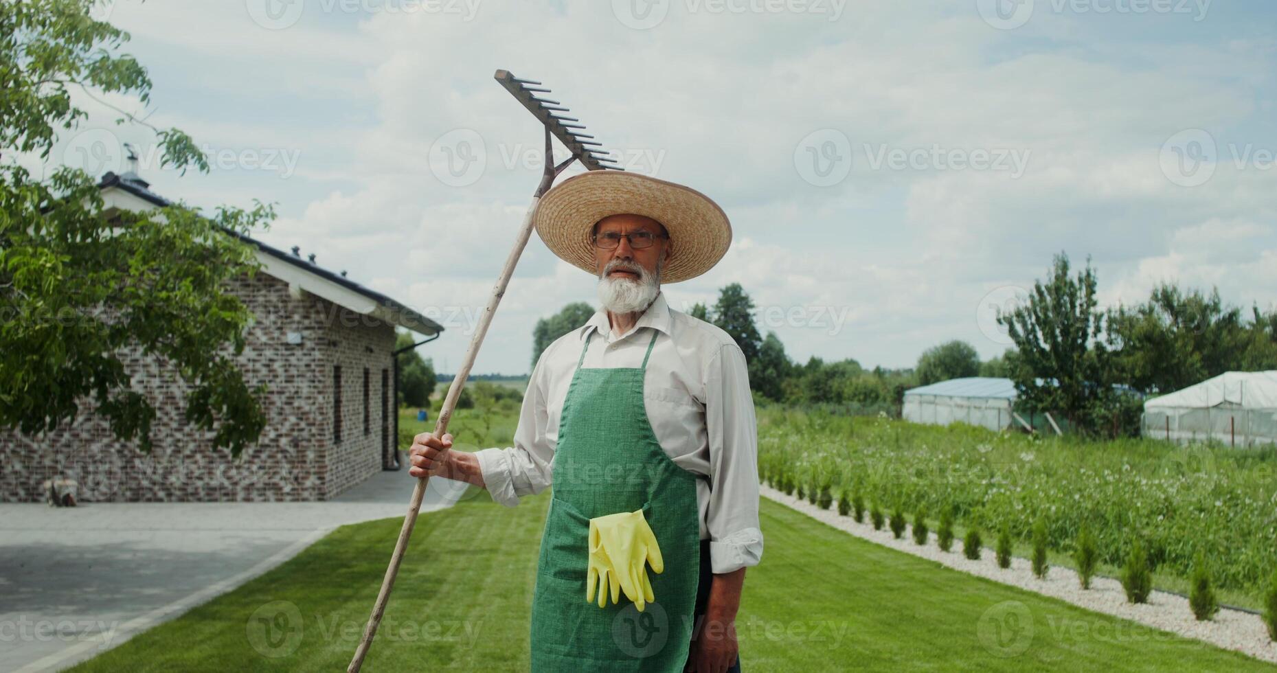 An elderly man with a gray beard, dressed in an apron and hat, smiles while looking at the camera standing in the backyard with a rake in his hand photo