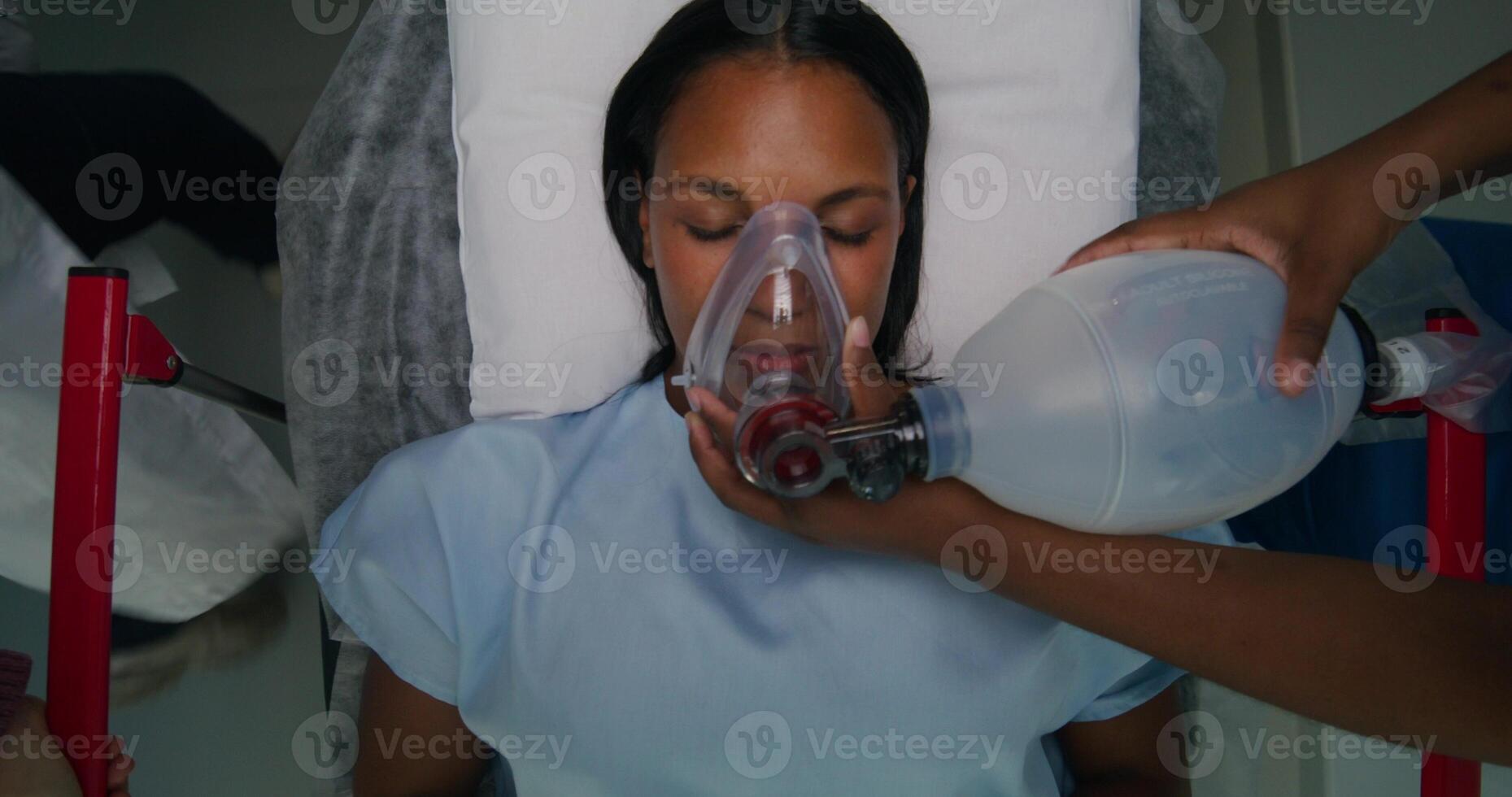 The patient is lying on a gurney with her eyes closed. The nurse gives her air through an oxygen mask. View from above. Emergency photo