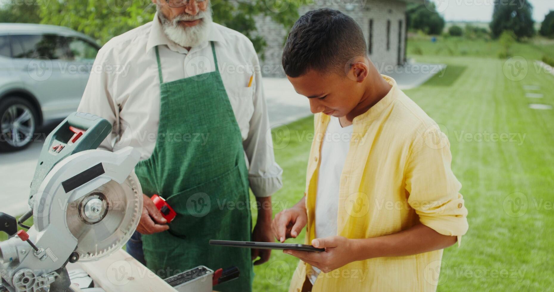 Grandfather and grandson are talking and using a tablet while standing in the backyard near a miter saw photo