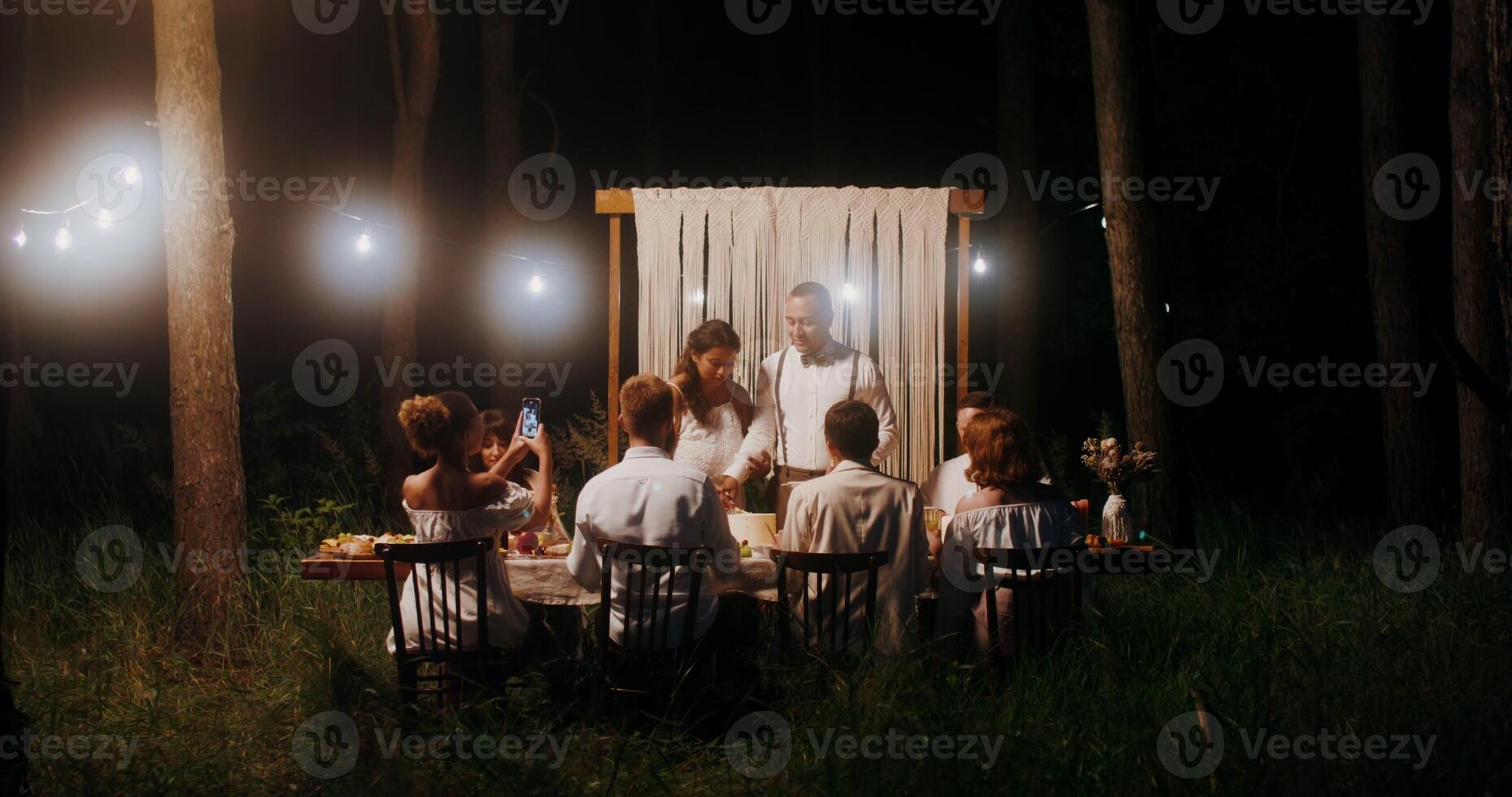 The bride and groom are cutting a wedding cake at an outdoor wedding feast. Guests take photos on their mobile phones - outdoor