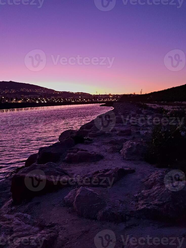 A valley bathed in the soft glow of post-sunset radiance emanates natural brilliance, as the horizon blazes with vivid purple and golden hues, while a mix of natural stones deepens the overall scene. photo