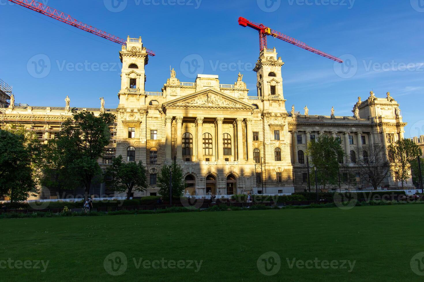 Front view of Museum of Ethnography in Budapest with neoclassical architecture, wide green lawn and construction cranes under clear blue sky photo