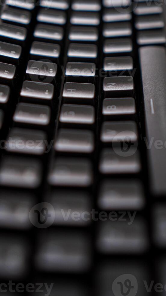 A close-up of a sleek black keyboard with square keys, resting on a dark, textured surface. The keyboard's sharp angles and minimalist design create photo