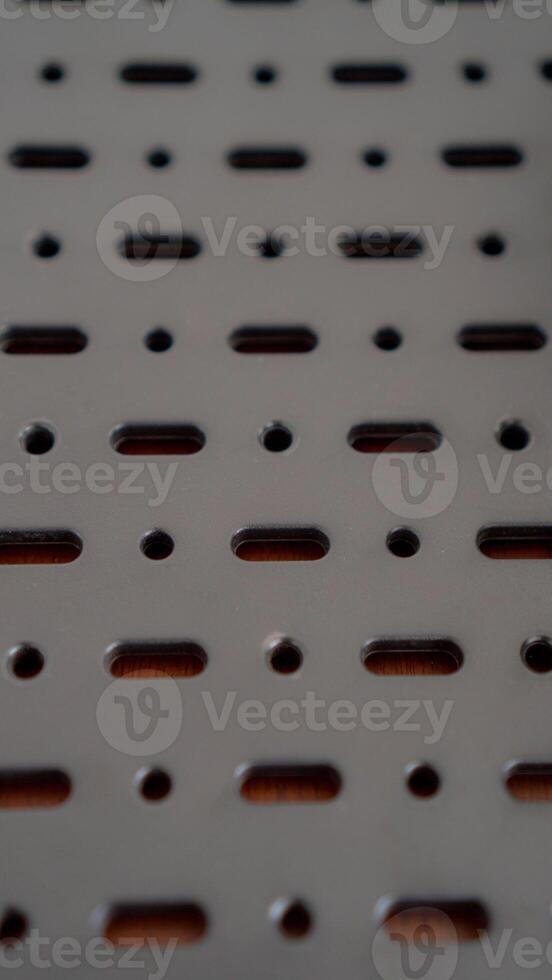 A close-up of a perforated metal surface with a repeating pattern of oval and circular holes. The dark, industrial aesthetic creates a sense of mystery and intrigue. The image highlights photo