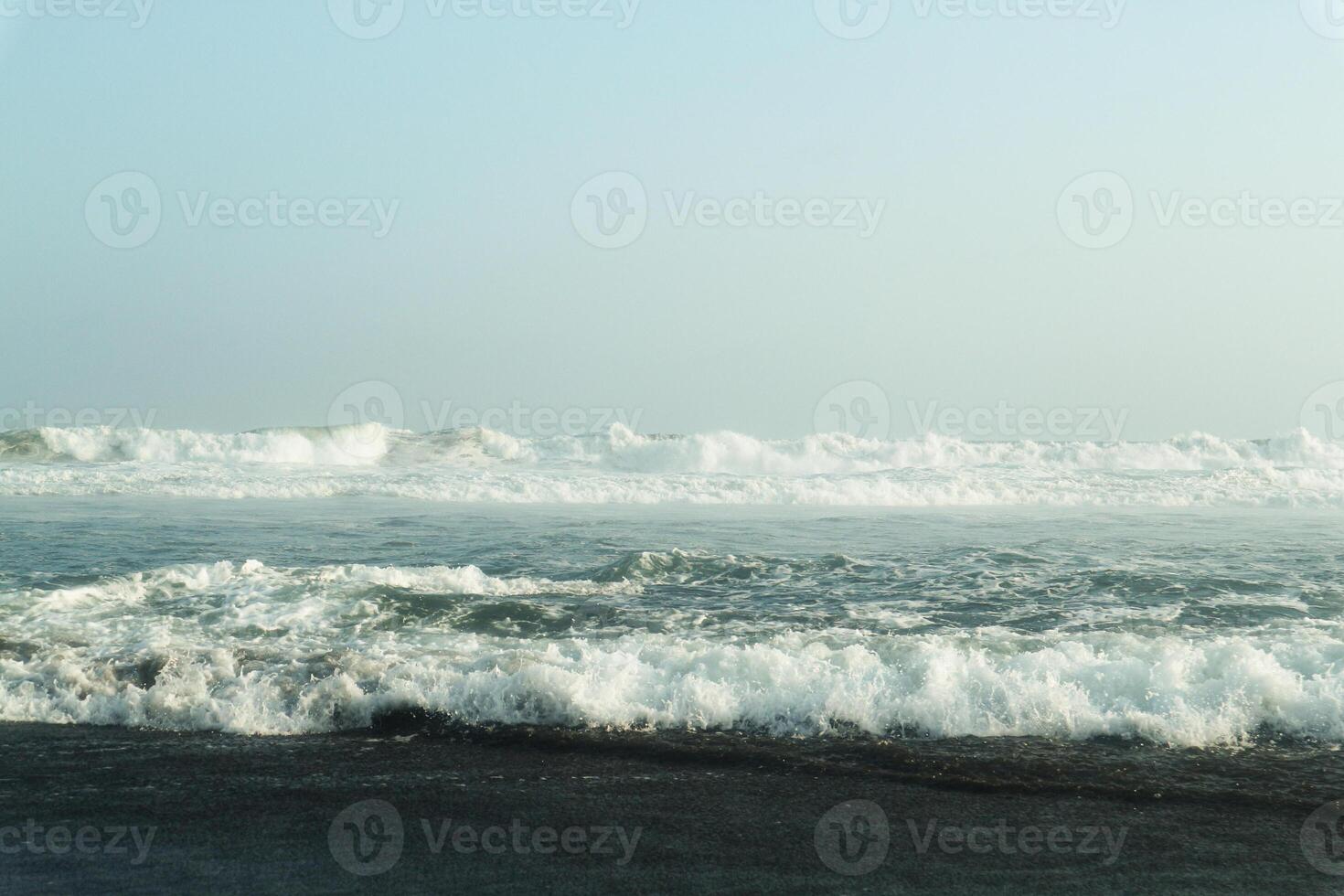 A serene coastal scene with powerful waves crashing onto a dark, sandy shore. The vast expanse of the ocean meets a clear, blue sky, creating a breathtaking panorama photo