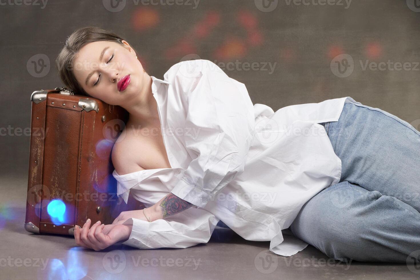 A beautiful girl with closed eyes lies on a vintage suitcase on a gray studio background with bright spots. The girl sleeps and dreams. photo