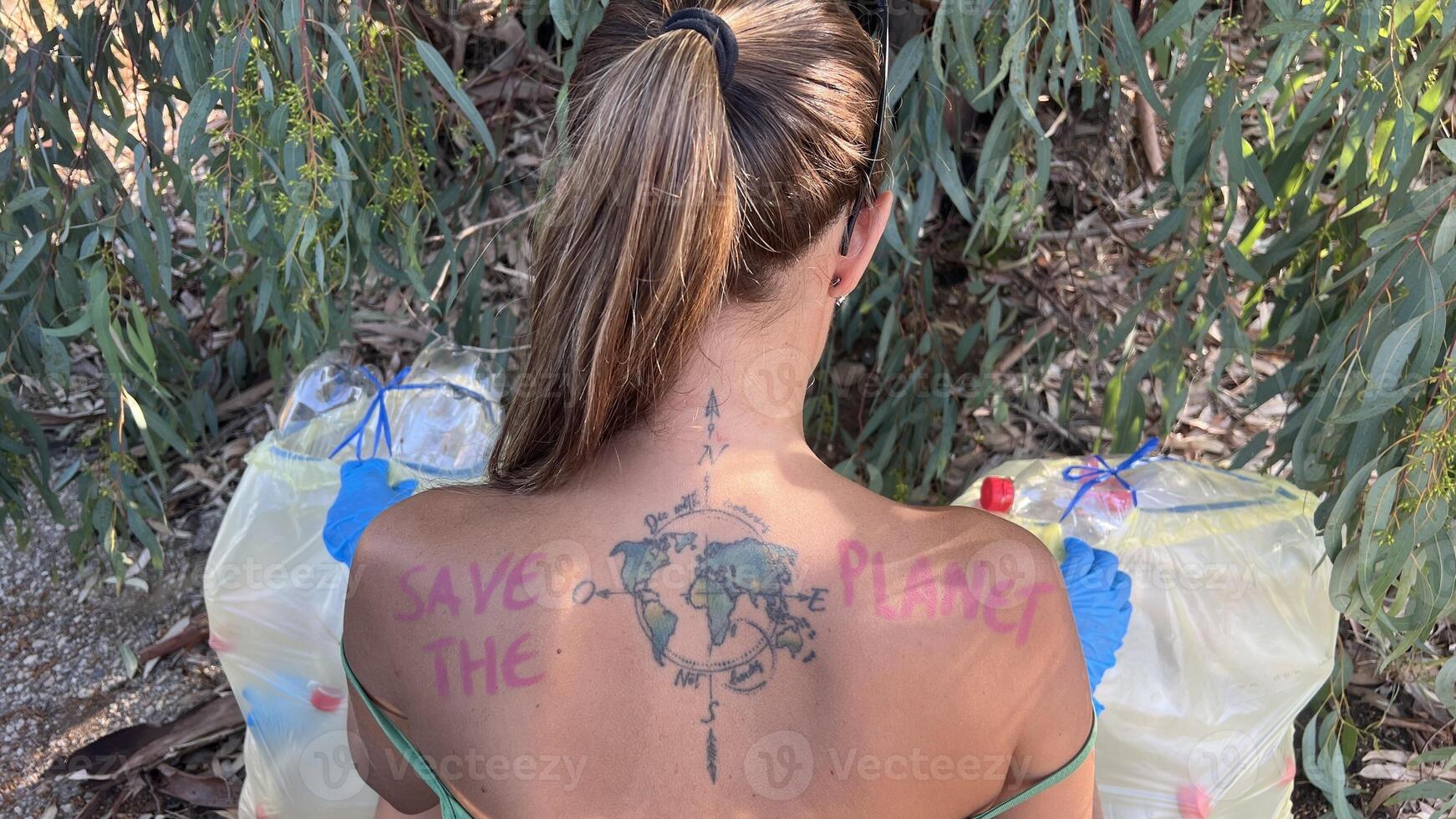 Woman with tattoo on her back that says SAVE THE PLANET and a map of the world. She is recycling plastic bottles discarded in the field. Concept of environment, responsibility, ecology, awareness photo