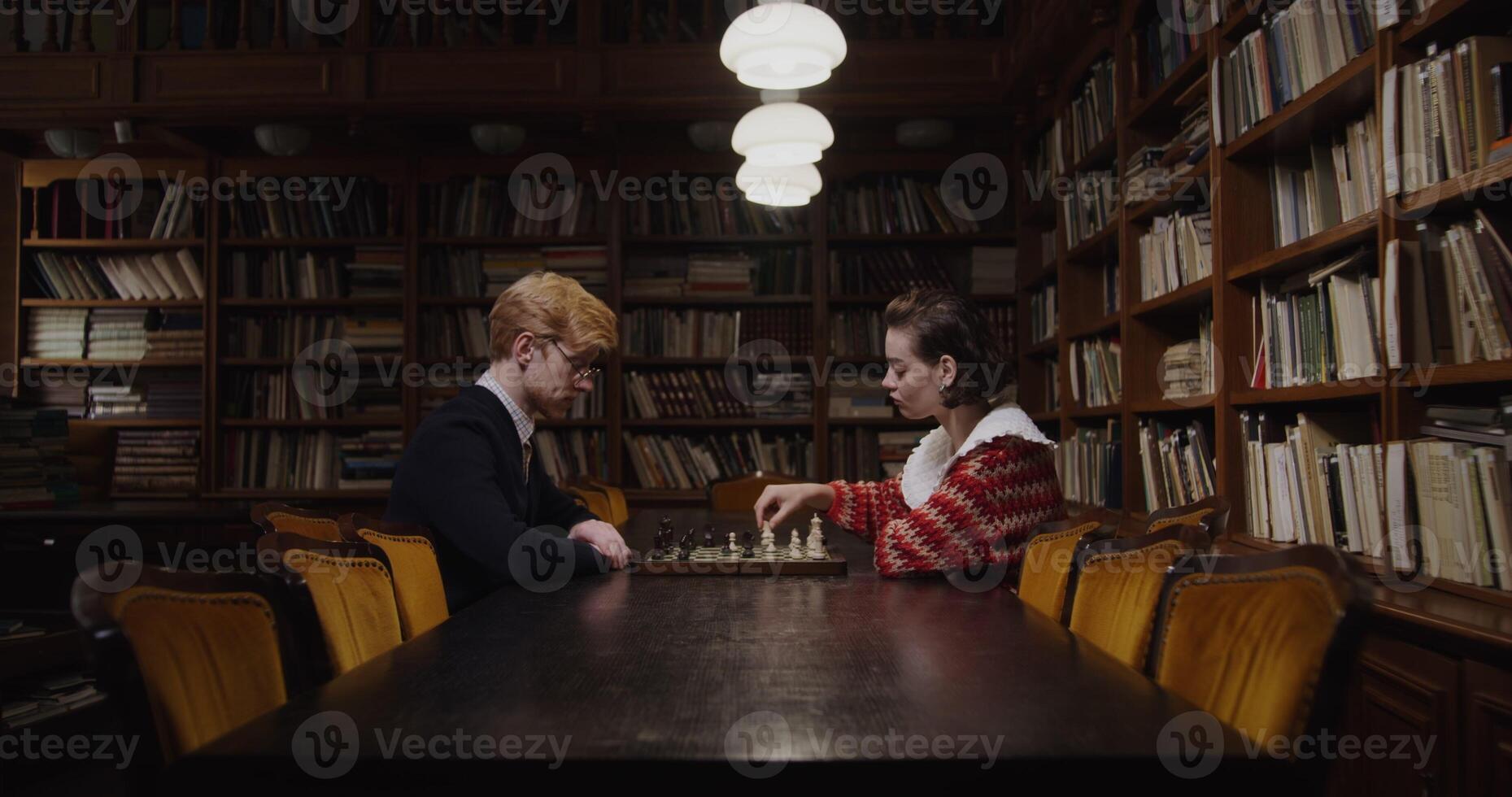 Thoughtful man and woman facing off in a chess game, seated in a peaceful library environment. photo
