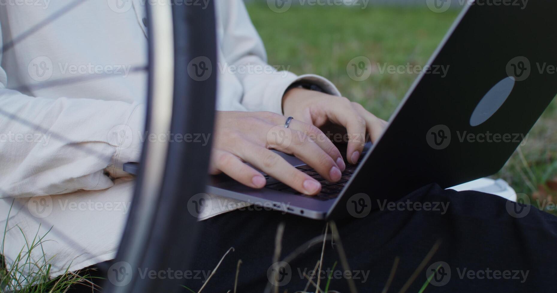 young man uses a laptop sitting on the grass under a tree outdoors, close-up. Bicycle Stands next to the man. Unidentified person, no face in the frame photo